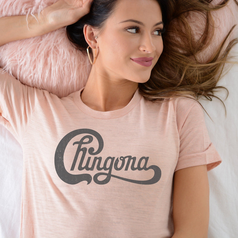 Woman laying in bed wearing peachy pink t-shirt with Chingona in dark gray cursive that looks like the Chicago logo