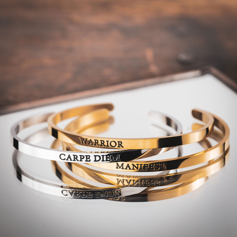 3 bangle cuff bracelets on a mirror. 1 gold bracelet says MANIFEST, 1 gold bracelet says WARRIOR, and 1 silver bracelet says CARPE DIEM 