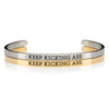 1 silver and 1 gold open cuff bracelet that says KEEP KICKING ASS in black lettering