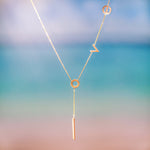 Gold lariat necklace with lowercase latters spelling "love". Necklace hanging in front of sunset