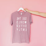 Hand holding a hanging pink T-shirt with reverse white letters that say See The World Differently backwards