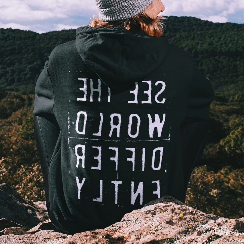 Sitting woman wearing black hooded sweatshirt with the words 'See The World Differently' printed backwards on the back in reverse white lettering 